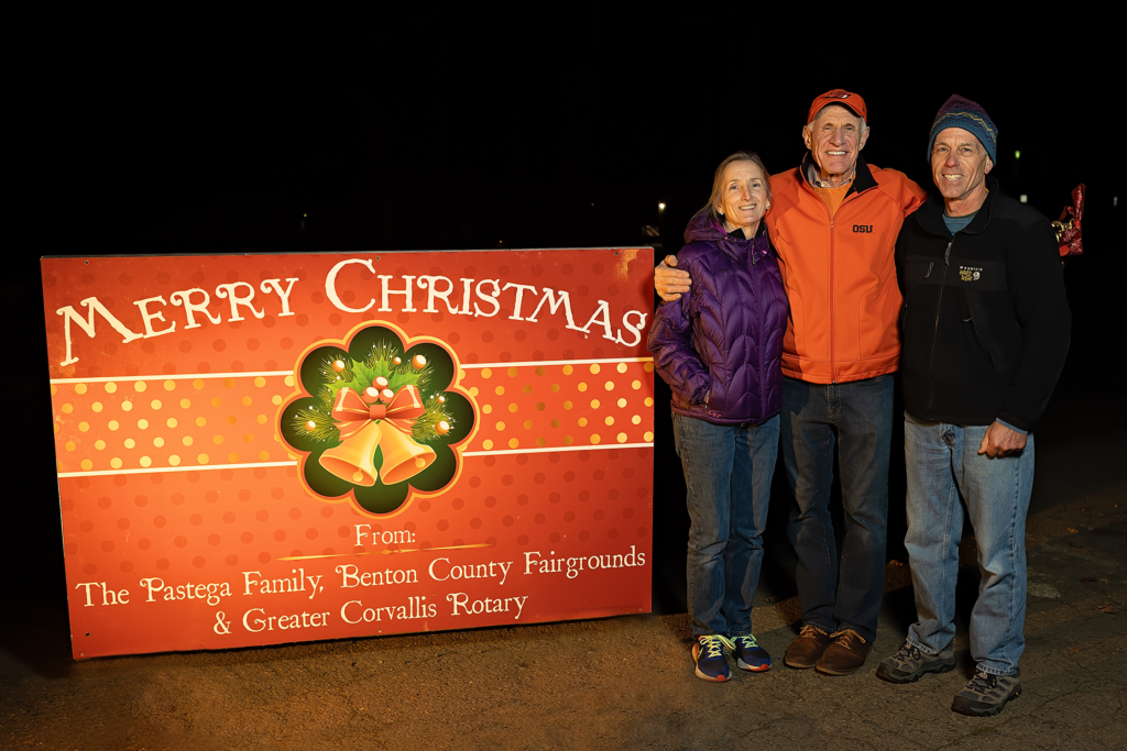 Ken Pastega and volunteers stand for a photo next to a red sign with the words "Merry Christmas from: the Pastega family, Benton County Fairgrounds, and Greater Corvallis Rotary."