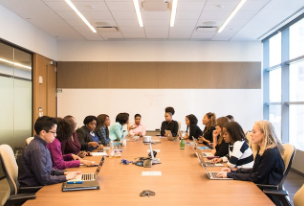 A room is filled with a long wooden table and a large group of people sit on both sides.